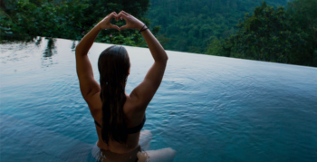 A woman performs yoga while sitting in an infinity pool looking over rainforest landscape